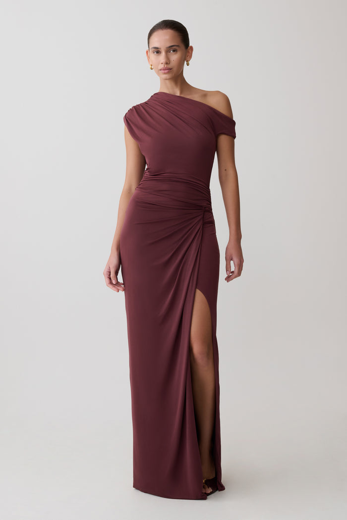 Woman wearing a burgundy off-shoulder gown with a slit on a light gray background